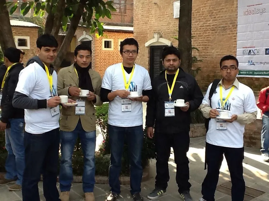 Five people in WordPress T-shirts and event lanyards holding cups of tea in a brick courtyard.