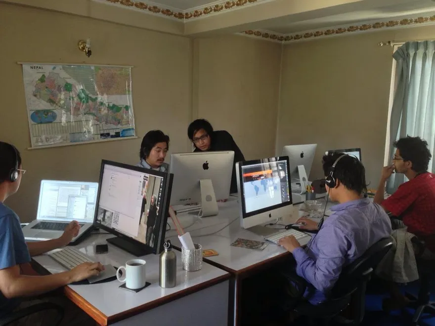 Five people working at iMacs and laptops around shared desks, with a map of Nepal on the wall.